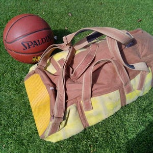 Könnte beinhalten: Eine braun-gelbe Sporttasche mit zwei Tragegriffen und einer Seitentasche, platziert neben einem orangefarbenen Spalding-Basketball. Die Tasche verfügt über ein strukturiertes gelbes Paneel an einem Ende und eine braune Stoffkonstruktion mit passenden Riemen.