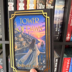 May include: A hardback book titled "Tower of Dawn" by Sarah J. Maas. The cover depicts a woman overlooking a city at sunset, with gold detailing. The book is held in front of a bookshelf filled with other books.