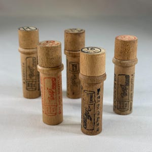 Könnte beinhalten: Eine Sammlung von fünf Vintage-Nadelbehältern aus Holz. Jeder Behälter hat einen zylindrischen Körper und einen Holzdeckel. Die Behälter sind mit Zahlen und Texten wie "Boye Brand" und "Buy New Shuttle" versehen. Die Behälter sind in verschiedenen Brauntönen gehalten.