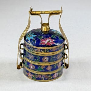 May include: A vintage, three-tiered, blue enamel container with a gold handle and accents. The container is decorated with pink, blue, and white floral designs. The container is likely used for food storage or display.