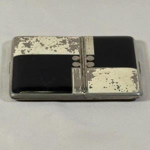 May include: Rectangular vintage cigarette case with a black and off-white geometric design. The case has a silver-colored metal frame and hinges. The off-white sections show signs of wear, adding to its antique charm.