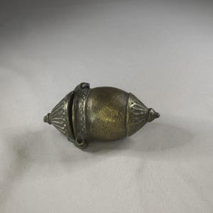 May include: A vintage brass container with a distinctive, symmetrical form. The container features a central, rounded body with conical ends and decorative, ridged detailing. A hinged section is visible, suggesting it opens. The brass has a warm, aged patina.
