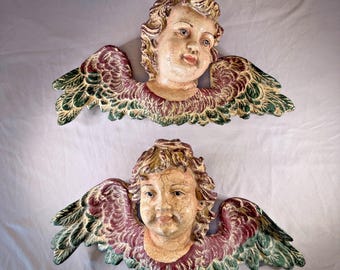 May include: Two antique cherub wall hangings. Each cherub has a cream-coloured face with curly hair and detailed wings. The wings are painted with burgundy, green, and gold accents. The cherubs have a distressed, aged appearance.