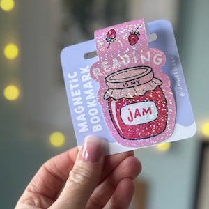 Peut inclure: Marque-page magnétique rose scintillant avec un motif de pot de confiture. Le texte « Reading is my Jam » est affiché, ainsi que deux illustrations de fraises et les mots « Magnetic Bookmark ».