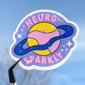 Puede incluir: Una pegatina morada y azul con un planeta rosa y amarillo con un anillo azul a su alrededor. El texto "NEURO SPARKLY" está en la pegatina.
