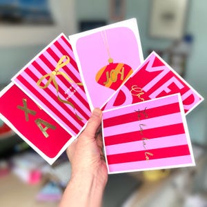 May include: A hand holds a collection of holiday greeting cards. The cards feature pink and red stripes, gold foil lettering, and festive graphics. Text includes "Happy Holidays," "Joy," and "Fa la la."