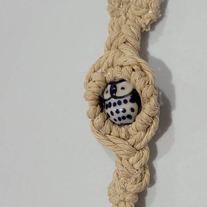 May include: A beige macrame keychain with a blue and white ceramic bead. The bead has a polka dot pattern. The keychain has a silver metal clasp.