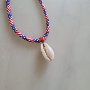 May include: A braided necklace with red, white, and blue cord. A white seashell pendant hangs from the necklace. The necklace is displayed on a light-colored surface.