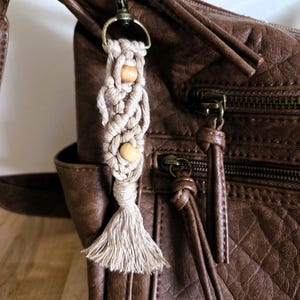 May include: A beige macrame keychain with wooden beads and a tassel, attached to a brown leather bag. The keychain is made of woven cord and has a natural, earthy aesthetic. The bag has a textured surface and metal hardware.