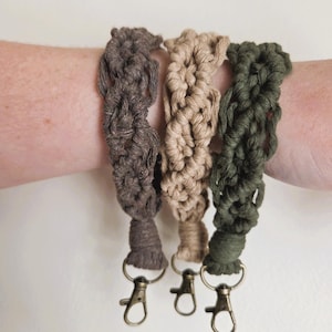 May include: Three macrame wristlets in shades of brown, tan, and olive green. Each wristlet features a woven design and a bronze-colored clasp. The wristlets are displayed on a person's wrist.