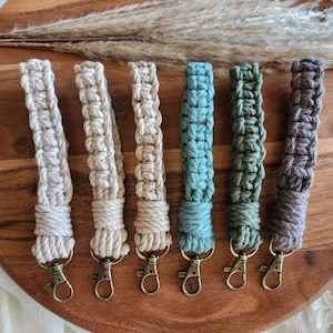May include: Seven handmade macrame keychains in various shades of beige, green, and brown. Each keychain features a metal key ring.