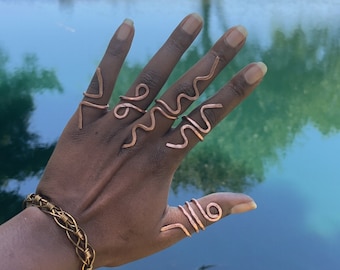 May include: A hand wearing multiple copper wire rings with wavy designs. The rings are worn on all fingers except the thumb. The hand is in front of a body of water with a green grassy area in the background.