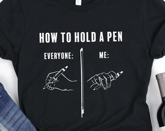 May include: Black t-shirt with white text that reads "How to hold a pen: Everyone: Me:" with a drawing of a hand holding a pen in two different ways.