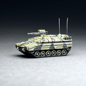 Puede incluir: Un vehículo blindado militar de camuflaje beige y negro con un pequeño cañón en la parte superior y dos antenas. El vehículo está sobre una superficie gris.