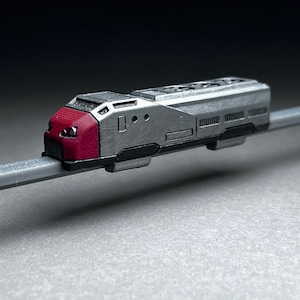 May include: A small, silver and red model train runs on a gray track. The train has a rounded front and a rectangular body with windows and details.