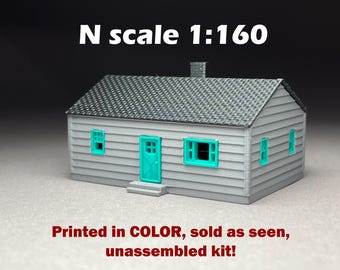 May include: A miniature model house in N scale 1:160, featuring a gray exterior with teal door and window frames. The roof is textured gray. The model includes a chimney and is an unassembled kit.