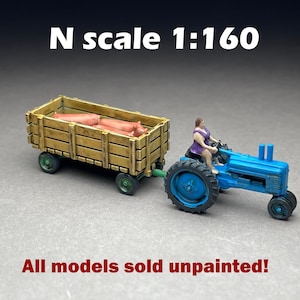 May include: A miniature blue tractor pulling a wooden cart filled with pink pigs. A figure in a purple top is driving the tractor. The text "N scale 1:160" and "All models sold unpainted!" are visible.