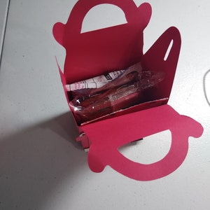 Puede incluir: Una caja de regalo rosa brillante, abierta, con asa, que contiene art&iacute;culos envueltos. La caja est&aacute; hecha de cartulina plegada y se encuentra sobre una superficie blanca. El asa es un recorte curvo en la parte superior.