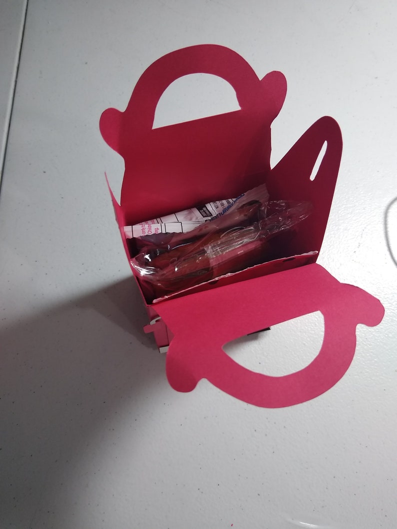 Puede incluir: Una caja de regalo rosa brillante, abierta, con asa, que contiene art&iacute;culos envueltos. La caja est&aacute; hecha de cartulina plegada y se encuentra sobre una superficie blanca. El asa es un recorte curvo en la parte superior.