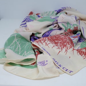 May include: A white silk scarf with a floral print in shades of green, red, and purple. The scarf has the text "Liberty of London Silk Cut" printed on it.