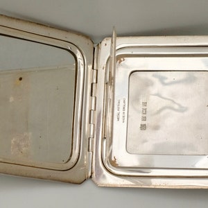 May include: A silver compact mirror with a hinged lid. The mirror is open, revealing a smaller mirror on the inside. The back of the compact is engraved with the text "MADE IN ENGLAND METAL FITTINGS GUARANTEED".
