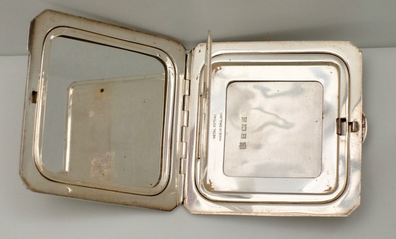 May include: A silver compact mirror with a hinged lid. The mirror is open, revealing a smaller mirror on the inside. The back of the compact is engraved with the text "MADE IN ENGLAND METAL FITTINGS GUARANTEED".