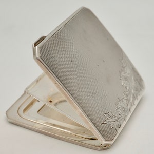 May include: A silver compact with a floral design engraved on the front. The compact is open, revealing a mirror inside.
