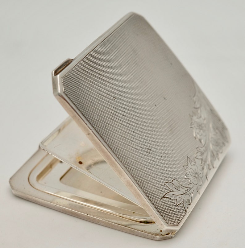 May include: A silver compact with a floral design engraved on the front. The compact is open, revealing a mirror inside.