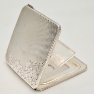 May include: A silver compact mirror with a floral design on the front. The mirror is open and the inside is a silver mirror.