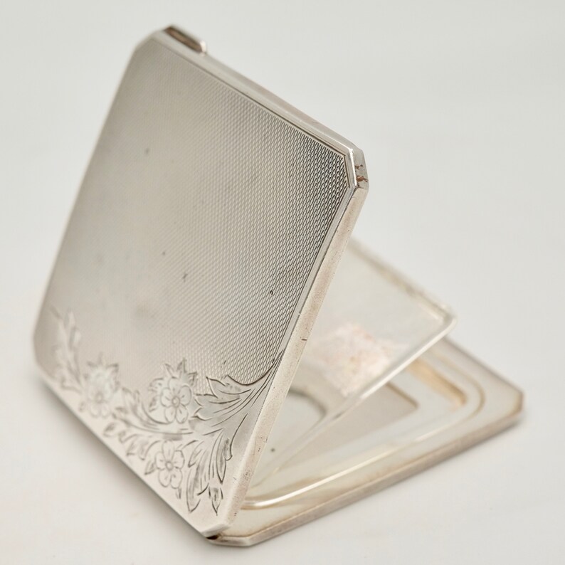 May include: A silver compact mirror with a floral design on the front. The mirror is open and the inside is a silver mirror.