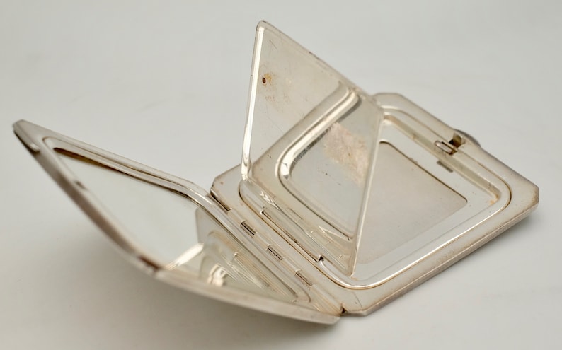 May include: A silver compact mirror with a hinged lid and two mirrors inside. The mirror is open and the lid is folded back.