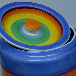 May include: A colorful ceramic jar with a lid. The jar is decorated with stripes of blue, orange, purple, and yellow. The lid has a rainbow design with a small orange knob in the center.