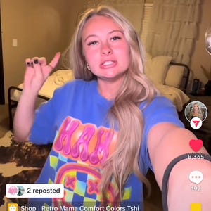 May include: A woman wearing a blue t-shirt with a pink, yellow, and green graphic design. The graphic design features a smiley face with a checkered pattern. The text "KAN" is visible on the shirt. The t-shirt is available for purchase from Retro Mama Comfort Colors Tshi.