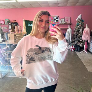 May include: White sweatshirt with a graphic print of a duck flying over a marsh. The sweatshirt has a crew neck and long sleeves.
