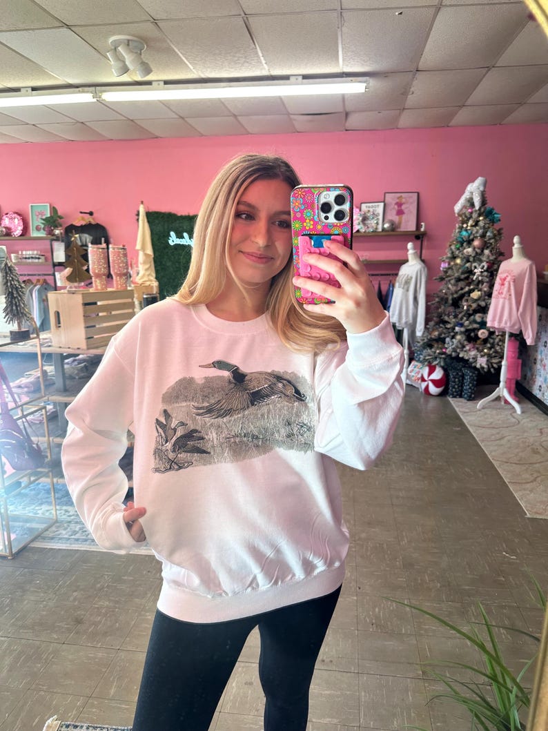 May include: White sweatshirt with a graphic print of a duck flying over a marsh. The sweatshirt has a crew neck and long sleeves.
