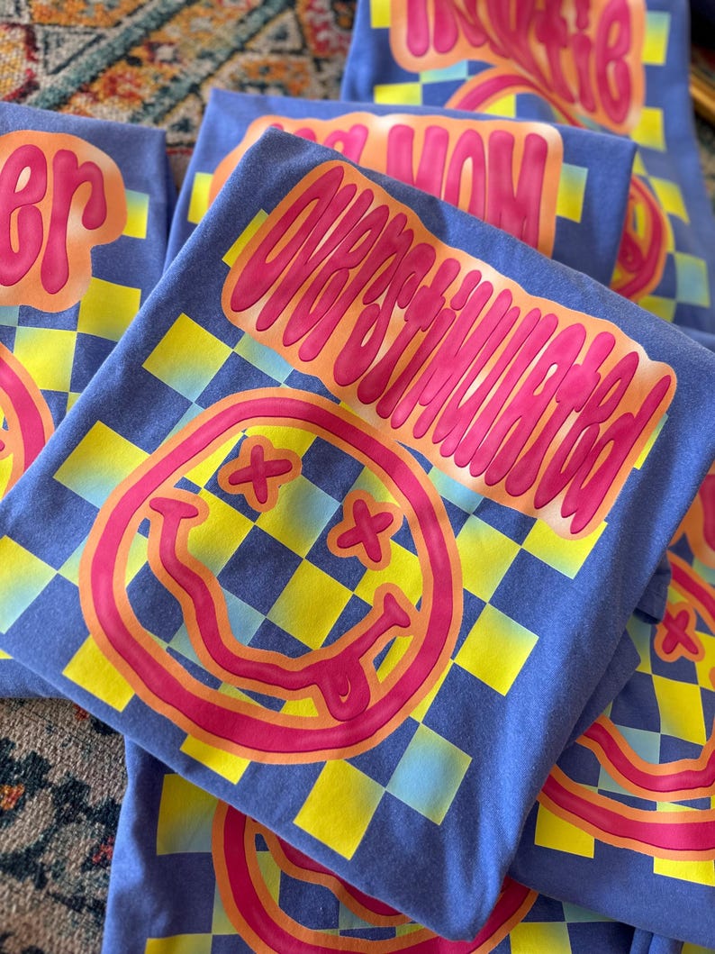 以下が含まれることがあります： 黄色と青のチェック柄の青いTシャツ。Tシャツには、舌を出したピンクのスマイリーフェイスと、ピンクの文字で「Overstimulated」と書かれています。