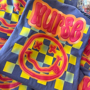 Puede incluir: Una camiseta azul con un patr&oacute;n a cuadros y una cara sonriente roja y amarilla. La palabra "NURSE" est&aacute; escrita en letras rojas y amarillas en la parte delantera de la camiseta.