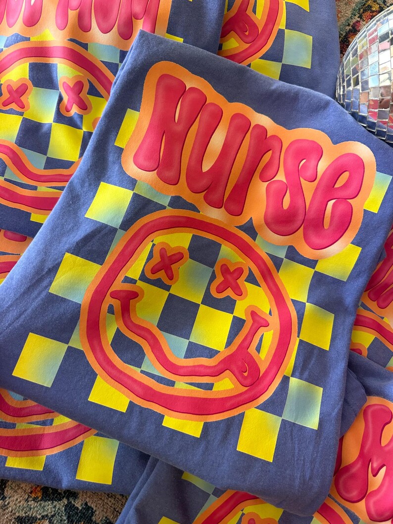 Puede incluir: Una camiseta azul con un patr&oacute;n a cuadros y una cara sonriente roja y amarilla. La palabra "NURSE" est&aacute; escrita en letras rojas y amarillas en la parte delantera de la camiseta.
