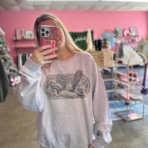 May include: A gray sweatshirt with a print of a flying duck in a camouflage pattern.