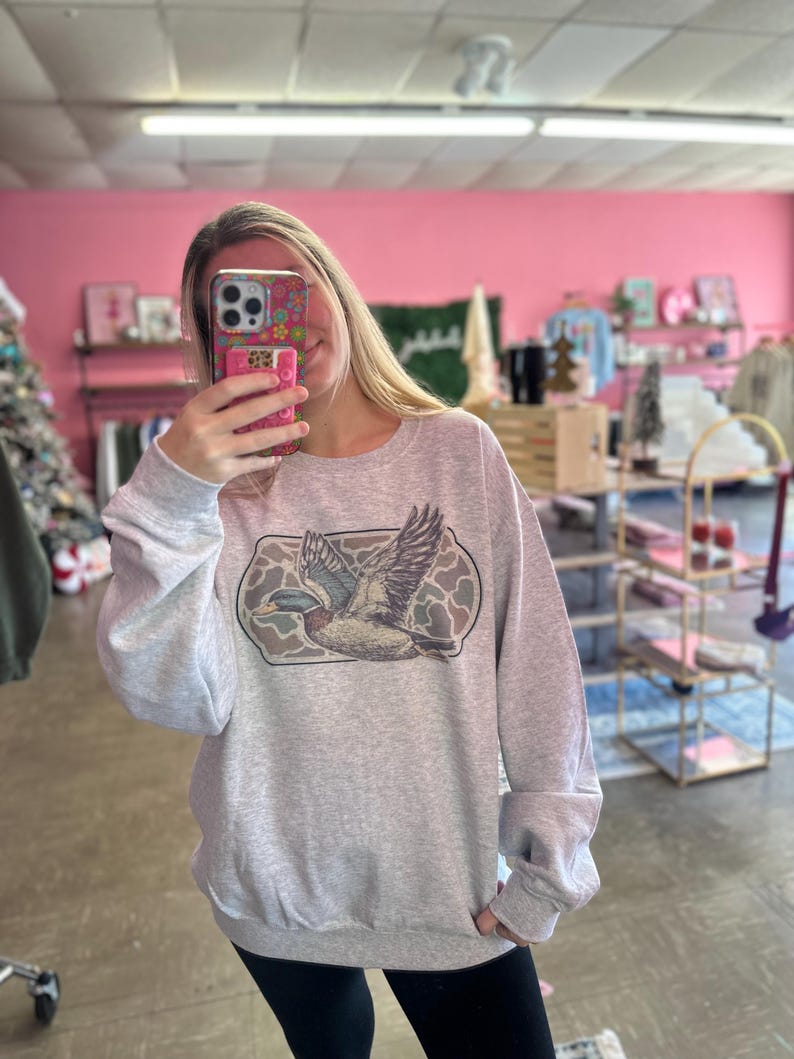 May include: A gray sweatshirt with a print of a flying duck in a camouflage pattern.
