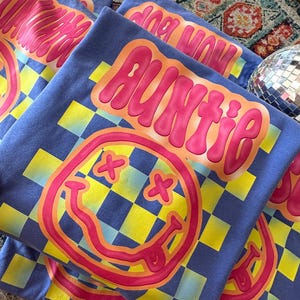 K&ouml;nnte beinhalten: Ein Stapel blauer T-Shirts mit einem gelben und blauen Karomuster, einem pinkfarbenen Smiley mit orangefarbenem Umriss und dem Wort "Auntie" in pinkfarbenen Buchstaben mit orangefarbenem Umriss.