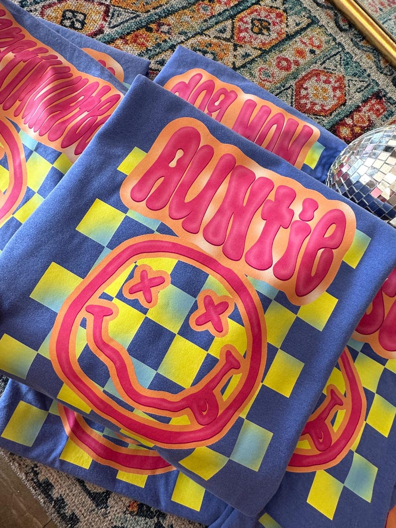 K&ouml;nnte beinhalten: Ein Stapel blauer T-Shirts mit einem gelben und blauen Karomuster, einem pinkfarbenen Smiley mit orangefarbenem Umriss und dem Wort "Auntie" in pinkfarbenen Buchstaben mit orangefarbenem Umriss.