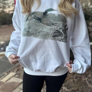 May include: White sweatshirt with a graphic of a mallard duck in flight. The duck is green, brown, and white. The background is a muted green and brown.