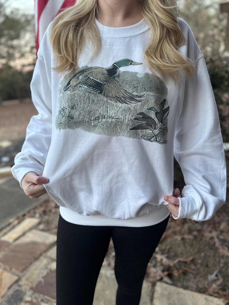 May include: White sweatshirt with a graphic of a mallard duck in flight. The duck is green, brown, and white. The background is a muted green and brown.