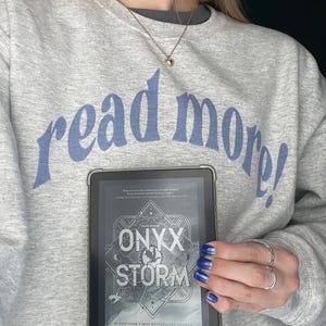 Puede incluir: Una persona sostiene un lector electr&oacute;nico Kindle que muestra la portada del libro "Onyx & Storm" de Rebecca Yarros. La portada del libro presenta un cielo oscuro y tormentoso con un dise&ntilde;o estilizado de una luna creciente y una estrella. El texto "Save the Dark" est&aacute; escrito sobre el t&iacute;tulo. El libro es un best-seller del New York Times. La persona lleva una sudadera gris con el texto "read more!" en azul.