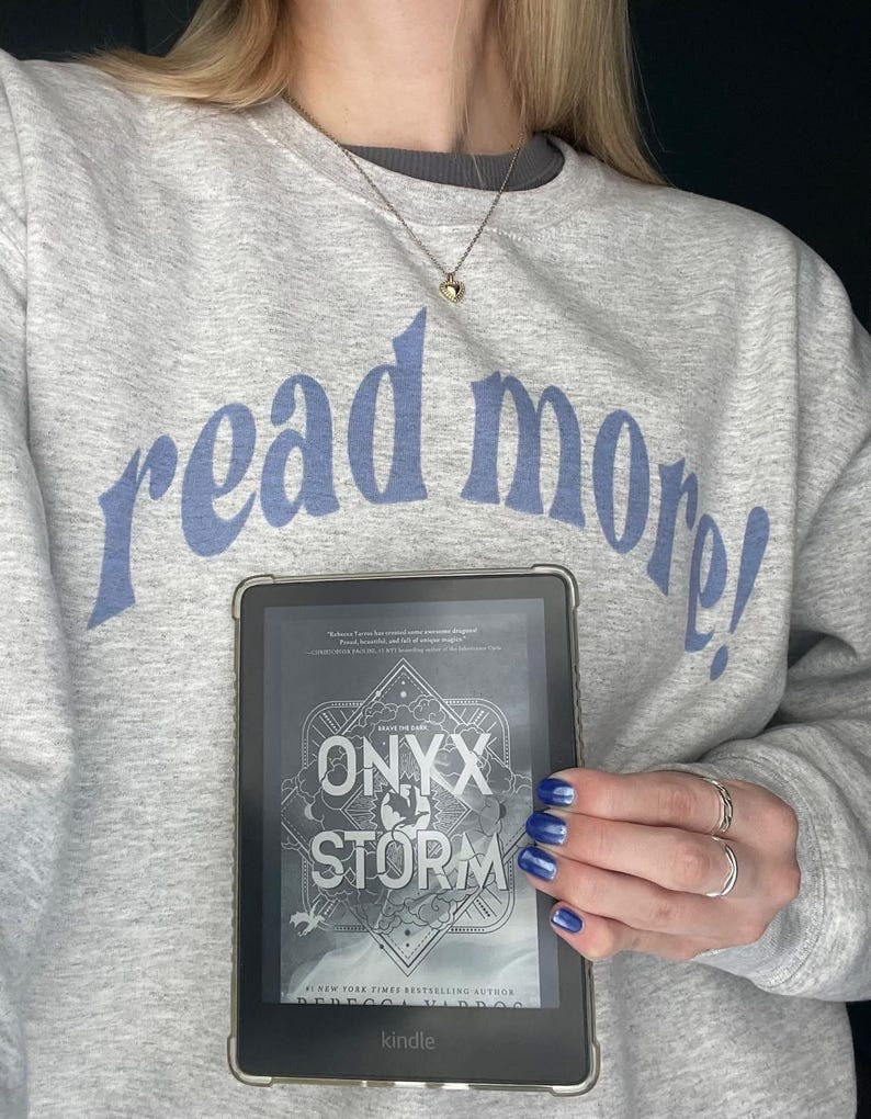Puede incluir: Una persona sostiene un lector electr&oacute;nico Kindle que muestra la portada del libro "Onyx & Storm" de Rebecca Yarros. La portada del libro presenta un cielo oscuro y tormentoso con un dise&ntilde;o estilizado de una luna creciente y una estrella. El texto "Save the Dark" est&aacute; escrito sobre el t&iacute;tulo. El libro es un best-seller del New York Times. La persona lleva una sudadera gris con el texto "read more!" en azul.