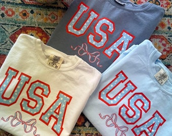 Può includere: Tre magliette con la scritta "USA" stampata in rosso e blu con stelle bianche. Le magliette sono di diversi colori: azzurro, bianco e celeste.