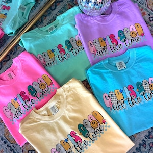 May include: A collection of colourful t-shirts with the phrase "God is good all the time" printed on them. The shirts are in various colours, including pink, blue, yellow, and green. Each shirt features a different design with a smiley face, stars, and lightning bolts.