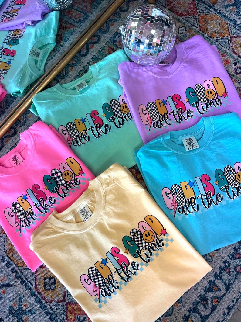 May include: A collection of colourful t-shirts with the phrase "God is good all the time" printed on them. The shirts are in various colours, including pink, blue, yellow, and green. Each shirt features a different design with a smiley face, stars, and lightning bolts.
