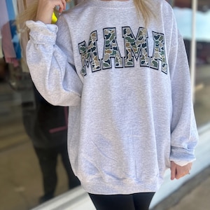 Puede incluir: Una sudadera gris con un gran gr&aacute;fico de "MAMA" en un patr&oacute;n de camuflaje. Las letras est&aacute;n delineadas en negro.
